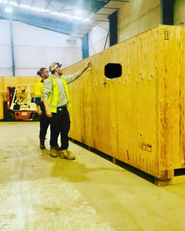 Two workers in safety vests inspect a large wooden packing crate inside a well-lit warehouse. One points at the crate, while a forklift stands in the background, highlighting an efficient industrial setting for wooden crates and export wooden pallets.