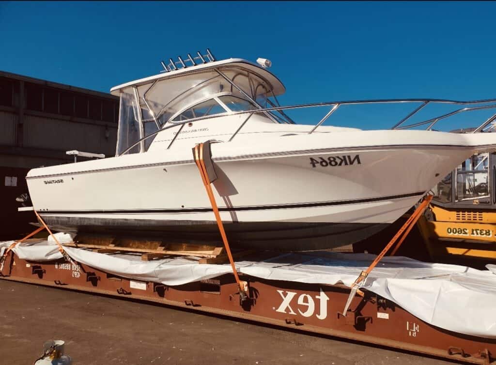 A white motorboat with a cabin is secured with orange straps on a flatbed shipping container beside wooden packing crates at an industrial dock, with a clear blue sky in the background.