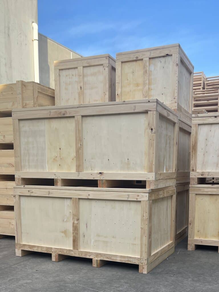 Several large wooden packing crates are stacked outdoors on a concrete surface, with a clear blue sky and part of a building visible in the background.