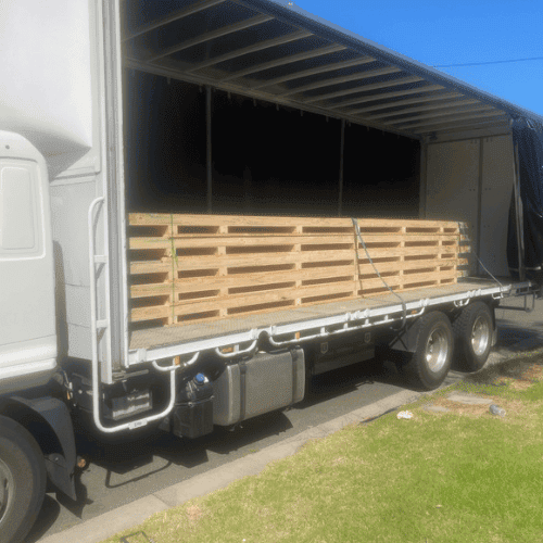 A flatbed truck with its side curtain open reveals a stack of export wooden pallets loaded and secured inside on a sunny day, parked next to a grassy area—ideal for reliable packaging solutions.