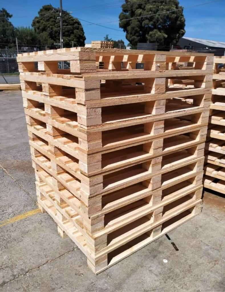 A stack of wooden pallets is neatly arranged outdoors on a concrete surface, with more pallets, wooden packing crates, and trees visible in the background under a clear blue sky.