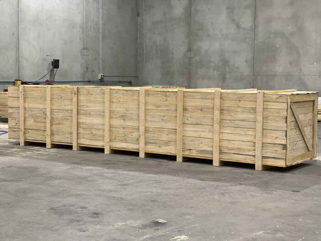 A large rectangular wooden packing crate with vertical supports sits on a concrete floor inside a spacious industrial warehouse with bare gray walls, ideal for export wooden pallets or on site packaging services.