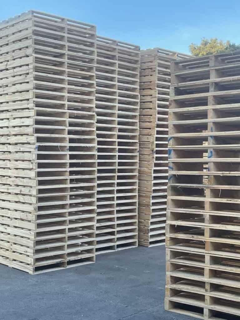 Tall stacks of wooden pallets and timber crates are arranged closely together outdoors on a paved surface, with a clear blue sky and some trees visible in the background—showcasing effective packaging solutions.